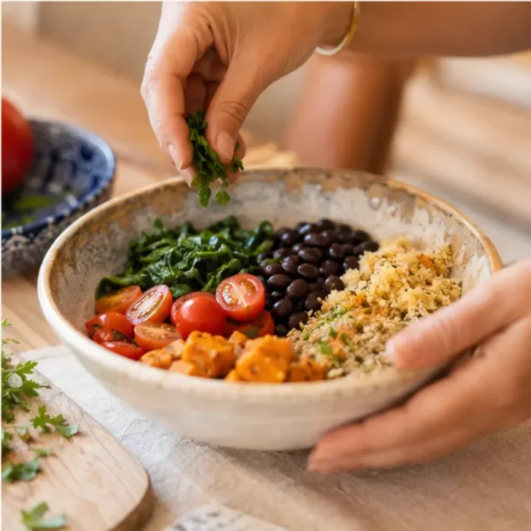 Mãos finalizando uma tigela de refeição colorida e equilibrada com feijão preto, cubos de abóbora assada, tomates cereja, verduras refogadas e cheiro-verde fresco